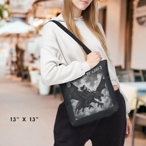 May include: A black tote bag measuring 13 inches by 13 inches, featuring a monochromatic graphic print of a figure riding a dragon amidst clouds. The text "GIRLS WILL BE GIRLS" is printed in white above the dragon illustration on the bag's surface.