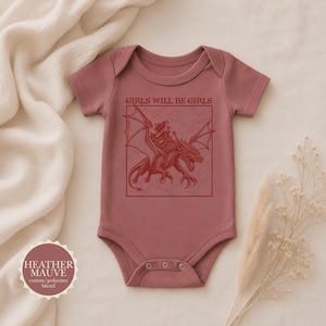 Puede incluir: Un body de beb&eacute; de manga corta en color malva jaspeado con un estampado gr&aacute;fico rojo oscuro de una persona montando un drag&oacute;n dentro de un marco cuadrado. Sobre la ilustraci&oacute;n, el texto dice GIRLS WILL BE GIRLS. Una insignia indica el color.