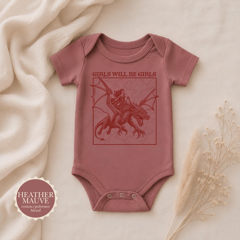Puede incluir: Un body de beb&eacute; de manga corta en color malva jaspeado con un estampado gr&aacute;fico rojo oscuro de una persona montando un drag&oacute;n dentro de un marco cuadrado. Sobre la ilustraci&oacute;n, el texto dice GIRLS WILL BE GIRLS. Una insignia indica el color.