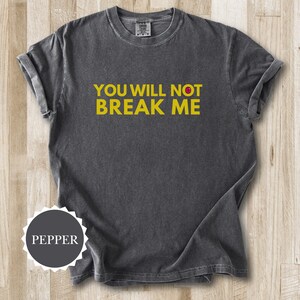 May include: A dark grey t-shirt with the yellow text "YOU WILL NOT BREAK ME". The shirt has a crew neck and short sleeves. The fabric looks soft and slightly faded, giving it a vintage appearance. The shirt is pepper-colored.