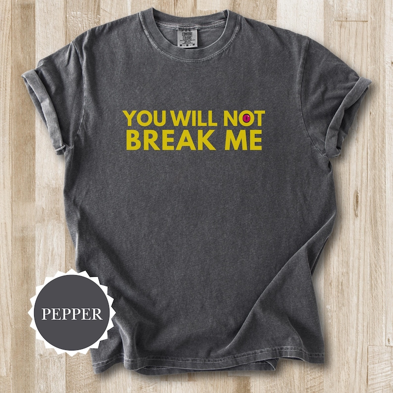 May include: A dark grey t-shirt with the yellow text "YOU WILL NOT BREAK ME". The shirt has a crew neck and short sleeves. The fabric looks soft and slightly faded, giving it a vintage appearance. The shirt is pepper-colored.