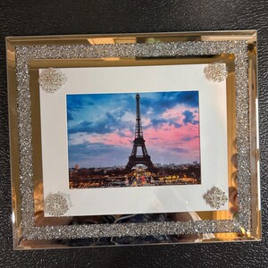 May include: A rectangular picture frame with a photograph of the Eiffel Tower at sunset. The frame has a gold border with a sparkling, glittery silver trim. Four decorative silver embellishments are on the frame.