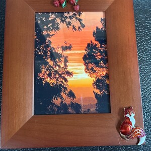May include: Wooden picture frame with a sunset landscape photo. The frame is decorated with a red and green jeweled strawberry branch and a jeweled fox. The frame has a warm, brown finish.