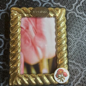 May include: A gold-colored picture frame with a twisted rope design. The word "dream" is above the photo area. A small, round emblem with a floral design is on the bottom right corner. The frame holds a photo with pink and white tones.