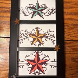 May include: Black framed wall art featuring three panels with star designs. Each panel displays a different colored star: teal, yellow, and red. Decorative scrollwork surrounds each star. Small star and bull head embellishments adorn the frame.