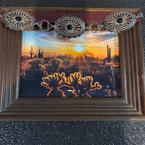 May include: A framed landscape photograph of a desert sunset with a decorative belt. The belt has three oval silver-toned medallions with black centers, connected by small rings. The frame is brown.