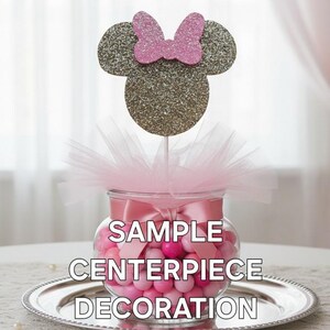 May include: A decorative centerpiece featuring a glass jar filled with pink candies, topped with a glittery mouse head and pink bow. The jar is adorned with a pink ribbon and tulle. Text on the jar reads "SAMPLE CENTERPIECE DECORATION."