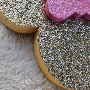 May include: Close-up of a glitter-covered Minnie Mouse head and bow. The ears are gold with iridescent glitter, and the bow is pink with glitter. The edges of the ears are a light brown color. The items are on a white, fluffy surface.