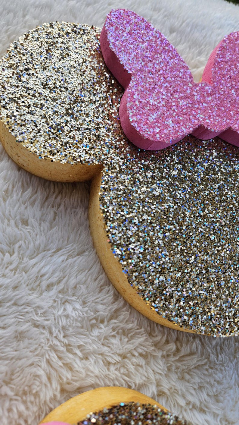 May include: Close-up of a glitter-covered Minnie Mouse head and bow. The ears are gold with iridescent glitter, and the bow is pink with glitter. The edges of the ears are a light brown color. The items are on a white, fluffy surface.