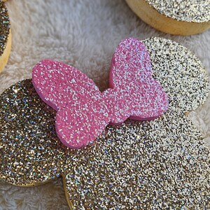 May include: Glittery gold and pink Minnie Mouse ear headbands. The ears are gold with glitter, and the bow is pink with glitter. The headbands are on a soft, white, fuzzy surface. The ears are a fun accessory.