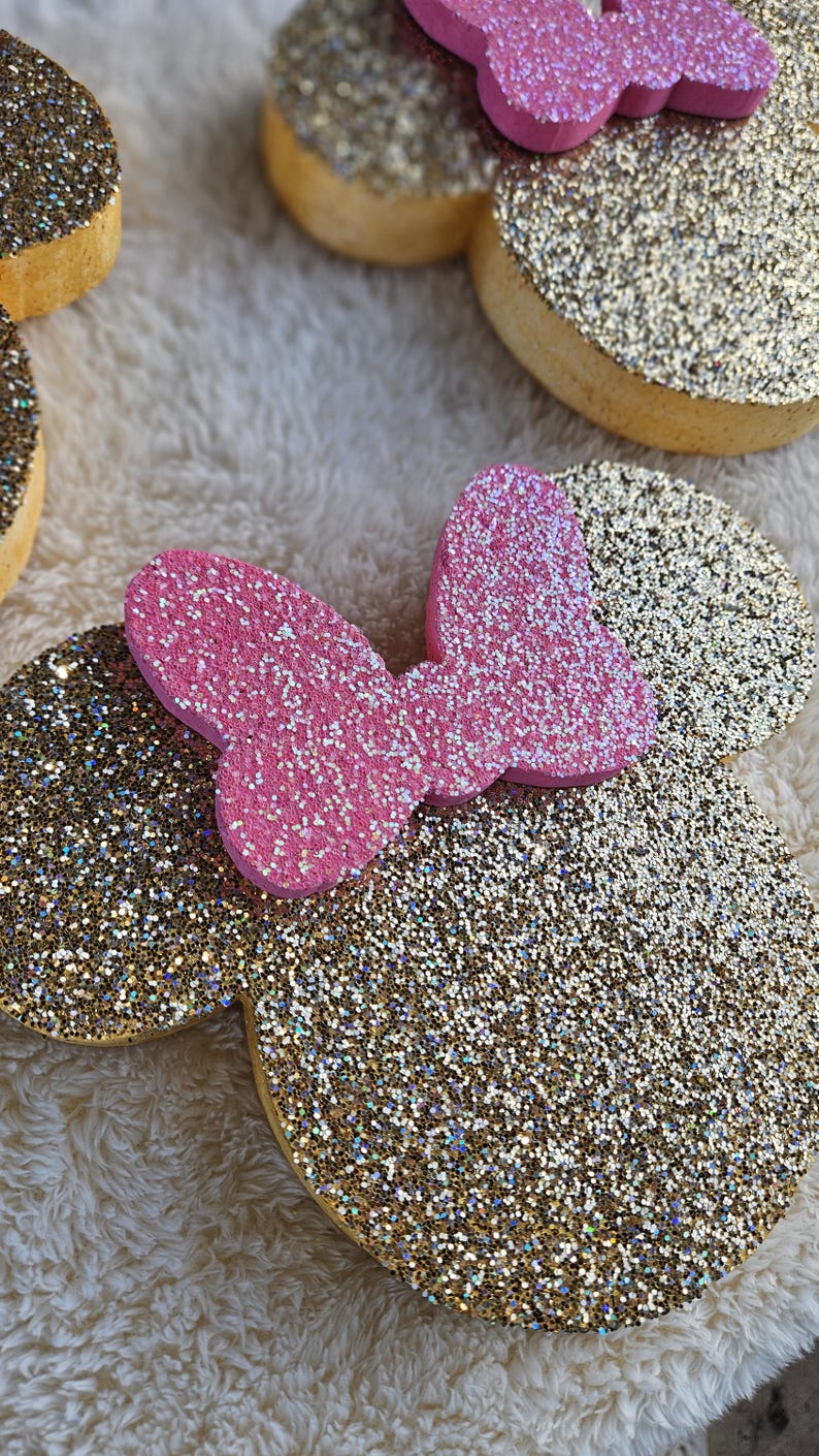 May include: Glittery gold and pink Minnie Mouse ear headbands. The ears are gold with glitter, and the bow is pink with glitter. The headbands are on a soft, white, fuzzy surface. The ears are a fun accessory.
