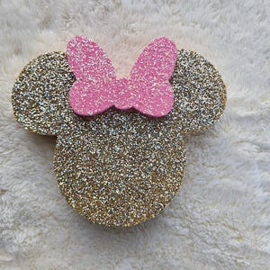 May include: A glittery gold Minnie Mouse head with a pink bow. The ears and head are covered in gold glitter, and the bow is a solid pink. The design is placed on a white, fluffy surface.