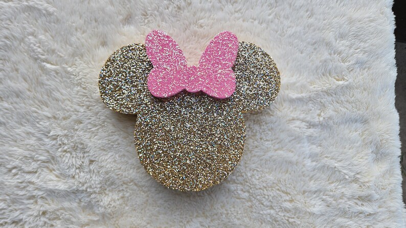 May include: A glittery gold Minnie Mouse head with a pink bow. The ears and head are covered in gold glitter, and the bow is a solid pink. The design is placed on a white, fluffy surface.