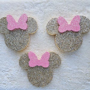 May include: Three glittery gold Minnie Mouse head shapes with pink bows. The shapes are arranged on a white, textured surface. The gold glitter catches the light, creating a sparkling effect. The pink bows add a pop of color.