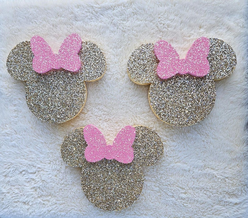 May include: Three glittery gold Minnie Mouse head shapes with pink bows. The shapes are arranged on a white, textured surface. The gold glitter catches the light, creating a sparkling effect. The pink bows add a pop of color.