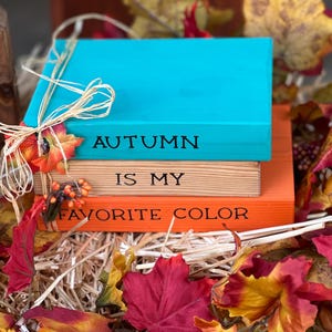 Peut inclure: Une pile de trois blocs de bois avec les phrases "AUTUMN IS MY FAVORITE COLOR" peintes dessus. Le bloc supérieur est turquoise, le milieu est en bois naturel et le bas est orange. Les blocs sont décorés de feuilles d'automne et de raphia.