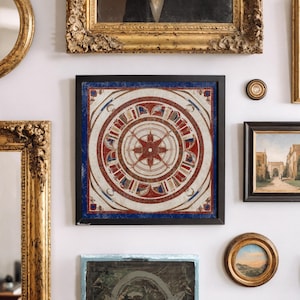 May include: Square framed artwork with a compass rose design in red, white, and blue. The artwork is surrounded by a black frame and is displayed on a white wall with other framed art pieces.