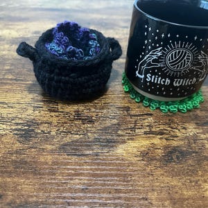 Puede incluir: Un caldero negro de ganchillo lleno de hilo morado y azul está junto a una taza negra. La taza tiene las palabras "Stitch Witch" y un diseño de ovillo de lana. Un posavasos verde de ganchillo está debajo de la taza.