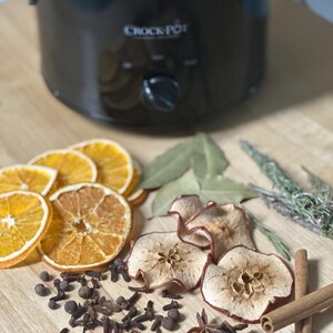 May include: A black Crock-Pot slow cooker with dried orange slices, apple slices, cloves, star anise, cinnamon sticks, and herbs arranged on a wooden surface. Ideal for making potpourri or simmering spices.