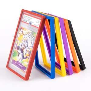 May include: A graded trading card in a red frame, leaning against a row of colorful frames. The card features a character illustration with Japanese text. The frames are in blue, yellow, purple, orange, and black.