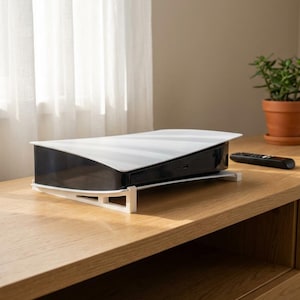 May include: A white and black gaming console on a white stand, with a black remote control. The console is on a light brown wooden table, near a window with white curtains and a potted plant.