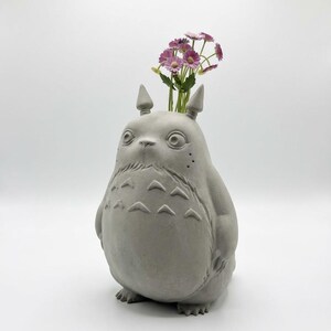 Puede incluir: Un jarrón de hormigón gris con forma de personaje de dibujos animados, con un cuerpo redondo, orejas pequeñas y una expresión amigable. El jarrón contiene un pequeño ramo de flores rosas y moradas, añadiendo un toque de color.