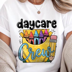 Può includere: Una t-shirt bianca con una stampa grafica di una scatola di pastelli gialla riempita con pastelli arancioni, rossi, blu, viola e rosa. Il testo "daycare" appare in nero sopra e la parola "Crew" è scritta in blu al di sotto.