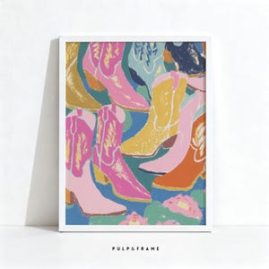 May include: A framed art print featuring a colorful arrangement of cowboy boots. The boots are in shades of pink, yellow, orange, and blue, with a white frame. The text "PULP & FRAME" is at the bottom.