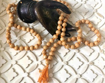 May include: A wooden mala bead necklace with a tassel, resting on a white surface with a decorative pattern. The necklace is wrapped around a bronze hand sculpture.