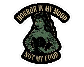 May include: A sticker with a green-skinned woman with black hair and tattoos, wearing a black top. The text "HORROR IN MY MOOD" curves above her, and "NOT MY FOOD" is below. The design has a vintage, spooky aesthetic.