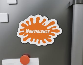 May include: A white-bordered magnet with an orange splatter design and the word "Nonviolence" in white. The magnet is affixed to a gray surface, along with other magnets and a paper. The design is reminiscent of a popular 90s television network.