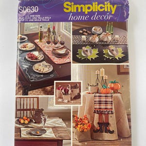 May include: A Simplicity home decor sewing pattern featuring five different table runner designs. The designs include a chalkboard table runner, a fall-themed table runner with pumpkins and boots, a table runner with a plaid design, a table runner with a leaf design, and a table runner with a geometric design.