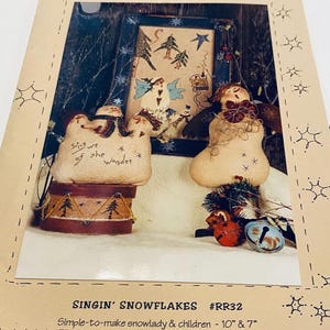 May include: A craft pattern titled "Singin' Snowflakes" by Reets' Rags To Stitches. The image features a framed stitchery with angels and trees, along with two snowlady figures and a drum. The pattern includes instructions for 10" and 7" snowlady figures.