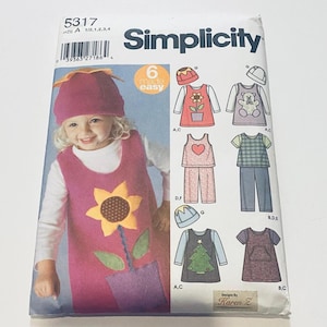 May include: A Simplicity 5317 sewing pattern envelope for children's clothing. The front features a photograph of a child wearing a pink jumper with a sunflower appliqué and a matching hat. Illustrations show variations including a bear, heart, and tree.