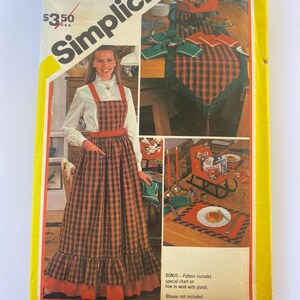 May include: A Simplicity sewing pattern package featuring a woman wearing a plaid apron dress. The package includes a bonus chart on how to work with plaids. The dress has a red belt and trim. The package also shows a table setting with plaid placemats.