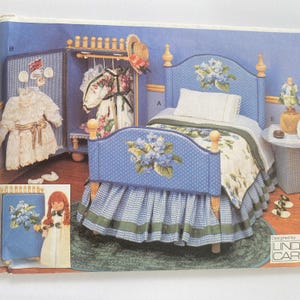 May include: A dollhouse bedroom set with a blue and white gingham bed, a white headboard with floral embroidery, a matching nightstand, and a doll-sized wardrobe with a white dress and a floral dress hanging inside. The set is designed by Linda Car.