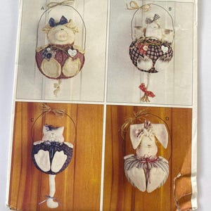 May include: Four handmade fabric animal ornaments, each suspended by a wire loop. The ornaments include a pig, a bird, a cat, and a bunny. Each animal has a decorative body and a bow. The background is a light beige color.