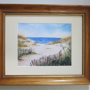 May include: A watercolor painting of a beach scene with a wooden fence leading to the ocean. The painting is framed in a light brown wood frame with a gold inner border.