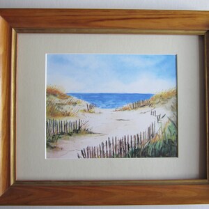 May include: A watercolor painting of a beach scene with a wooden fence leading to the ocean. The sky is blue with white clouds and the water is a deep blue. The sand is white and there are some green plants in the foreground.