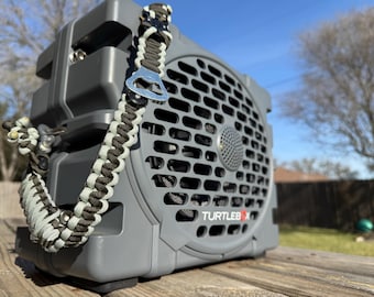 May include: A grey Turtlebox portable speaker featuring a custom paracord handle in olive green and light grey. The handle includes a silver metal bottle opener attachment. The speaker has a prominent circular grille and the brand name printed in white.