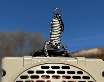 May include: A close-up of a pet carrier with a woven handle. The handle is made of black and white cord, attached to a metal ring. The carrier is beige with black vent holes, set against a blue sky.