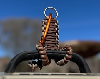 May include: A brown and tan paracord keychain with an orange carabiner clip and a silver ring. The paracord is wrapped around a black handle, set against a blurred blue sky background.