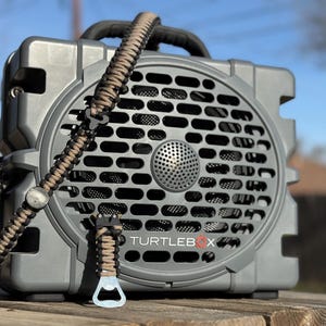 May include: A gray portable speaker with a black handle and a woven strap. The speaker has a circular grill and the text "TURTLEBOX". A bottle opener is attached to the strap. The speaker is sitting on a wooden surface.
