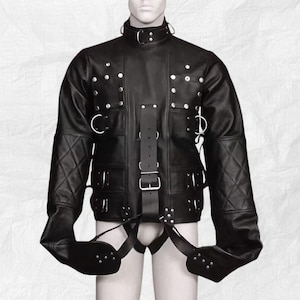 Puede incluir: Chaqueta de cuero negro con cuello alto, hombros con costuras de diamante y múltiples remaches y anillas en D de metal. La chaqueta presenta una correa central con hebilla y correas que se extienden a la parte inferior del cuerpo.