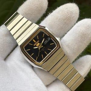 May include: A Seiko 5 automatic wristwatch with a black square dial, gold-tone hour markers, and gold hands. The watch features a day-date window at the three o'clock position and a silver-tone stainless steel link bracelet with a brushed finish.