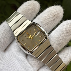 Puede incluir: Un reloj de pulsera automático Seiko 5 de tono plateado con caja rectangular y brazalete de eslabones integrados a juego. El reloj presenta una esfera marrón claro con marcadores de hora dorados, manecillas doradas y ventana de día y fecha.