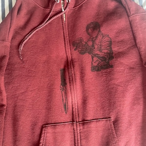 Könnte beinhalten: Burgunderfarbener Kapuzenpullover mit Reißverschluss, Vordertasche und Kapuze mit Kordelzug. Der Kapuzenpullover hat schwarze Grafiken einer Person mit einer Waffe, einem Dolch und einem Teddybär. Der Reißverschluss verläuft in der Mitte des Hoodies.
