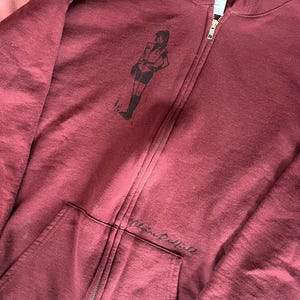 Könnte beinhalten: Burgunderfarbener Kapuzenpullover mit Reißverschluss und einer schwarzen Grafik einer Person auf der oberen linken Brust. Der Hoodie hat eine Kapuze mit Kordelzug, eine Vordertasche und den Namen "Claire Redfield" auf der unteren rechten Tasche.