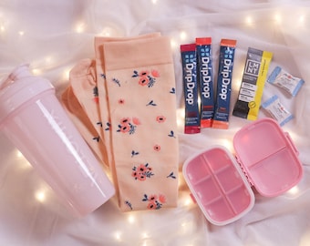May include: A collection of pink items, including a shaker bottle, floral socks, a pill organizer, and electrolyte packets. The socks have a peach background with pink and blue floral patterns. The items are arranged on a white surface.