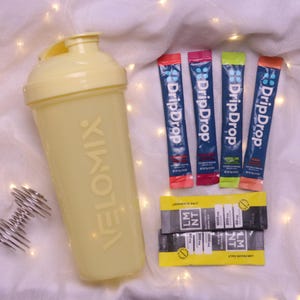 May include: A light yellow shaker bottle with the word VELOMIX printed on the side, a metal whisk, and several packets of electrolyte powder. The packets are labeled "DripDrop" and "LMNT". The background is a white cloth.